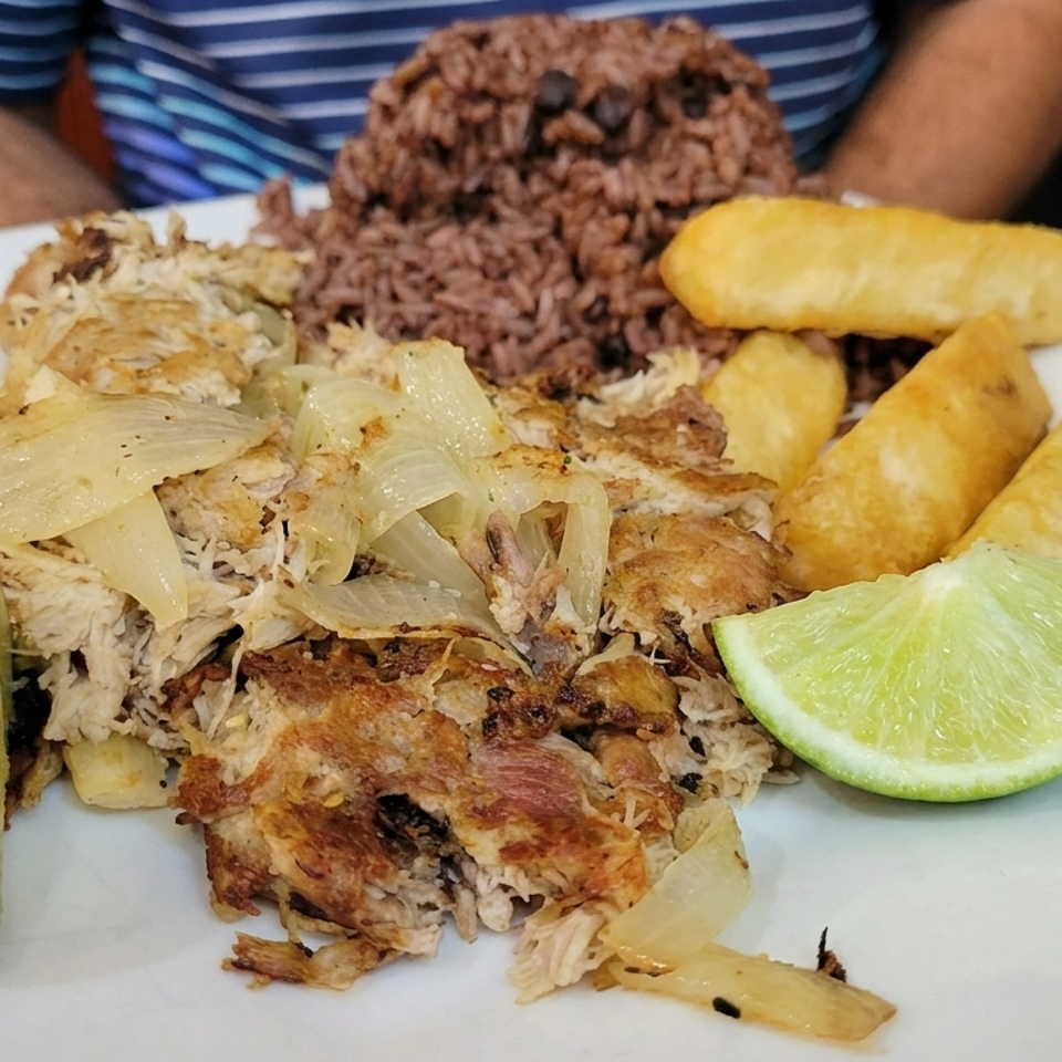 Plate of roasted shredded meat topped with onions, black beans and rice, fried plantains, and lime wedges.