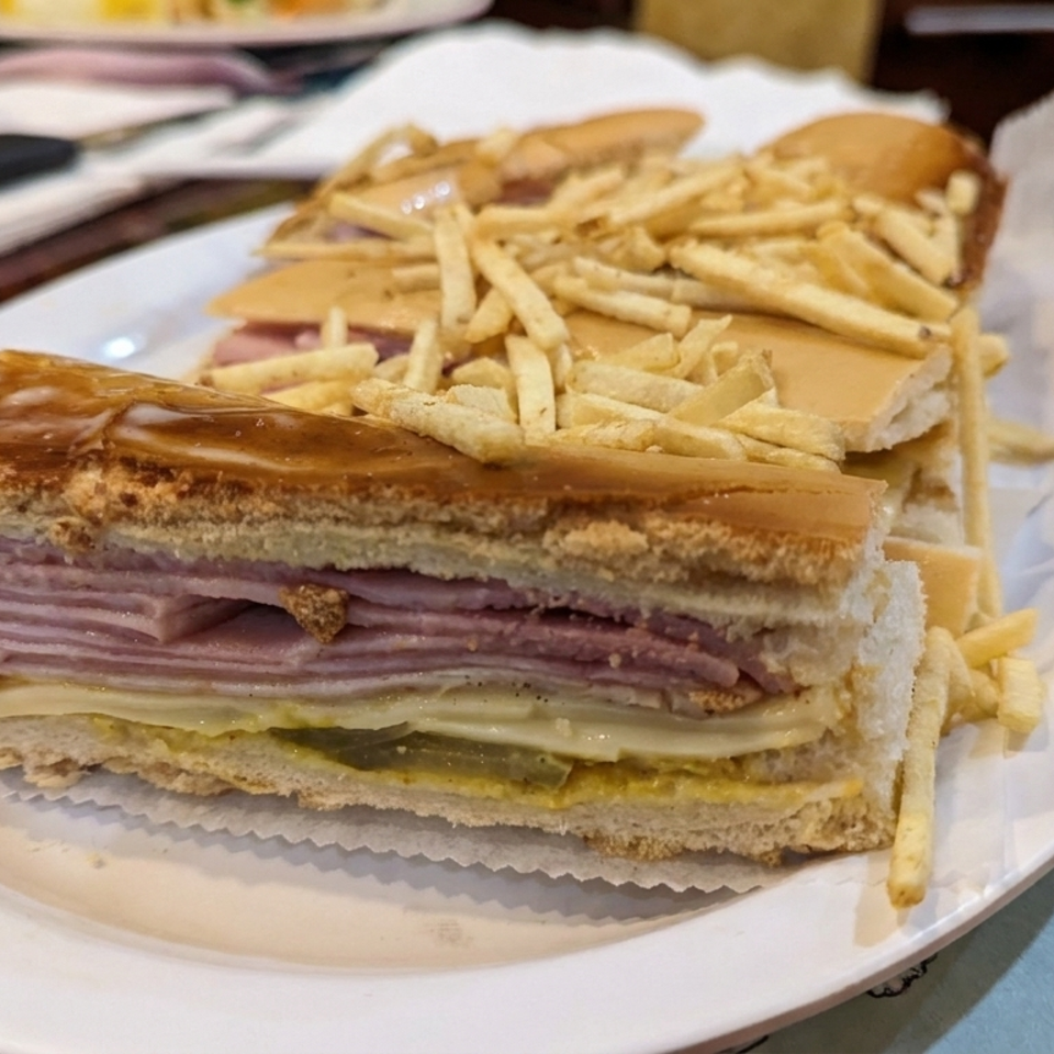 Layered ham and cheese sandwich served with thin French fries on a white plate at a restaurant table.